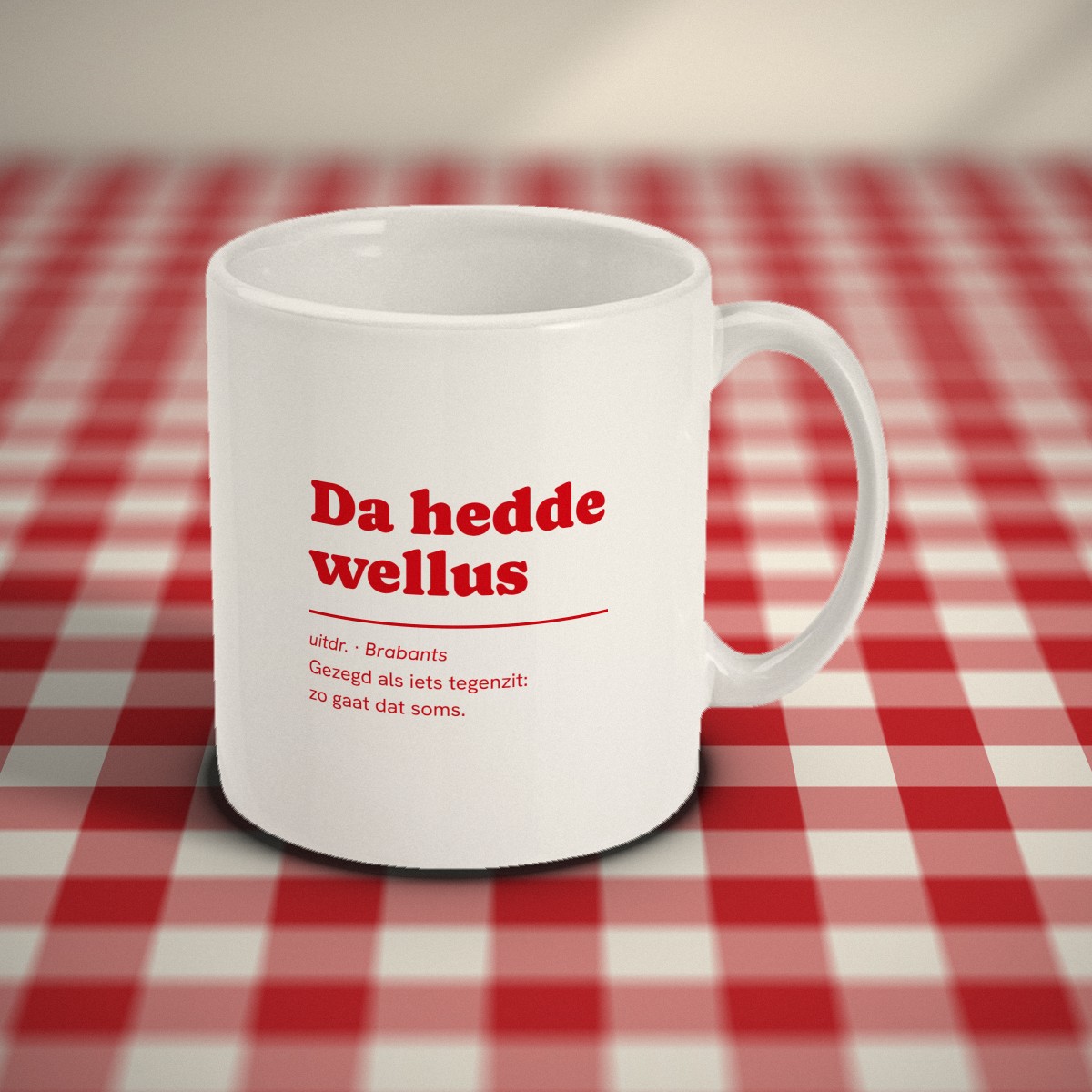 Voorkant: witte mok met in rode letters „Da hedde wellus” en de uitleg „Gezegd als iets tegenzit: zo gaat dat soms”, op een rood-wit geruit tafelkleed