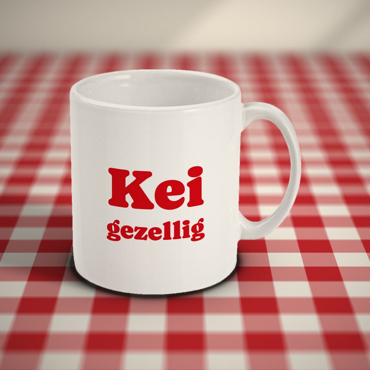 Voorkant: witte mok met in rode letters „Kei gezellig”, op een rood-wit geruit tafelkleed