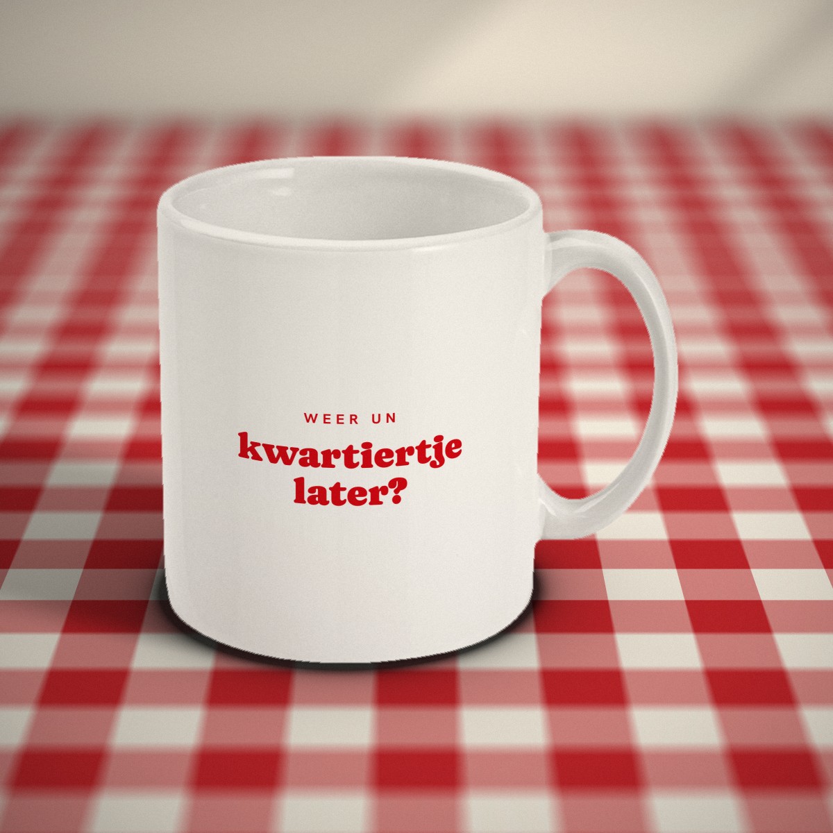 Voorkant: witte mok met in rode letters „weer un kwartiertje later?”, op een rood-wit geruit tafelkleed