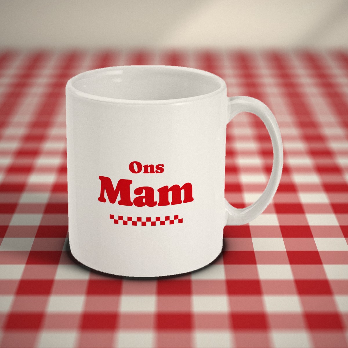 Voorkant: witte mok met in rode letters „Ons Mam” en een geblokt randje, op een rood-wit geruit tafelkleed