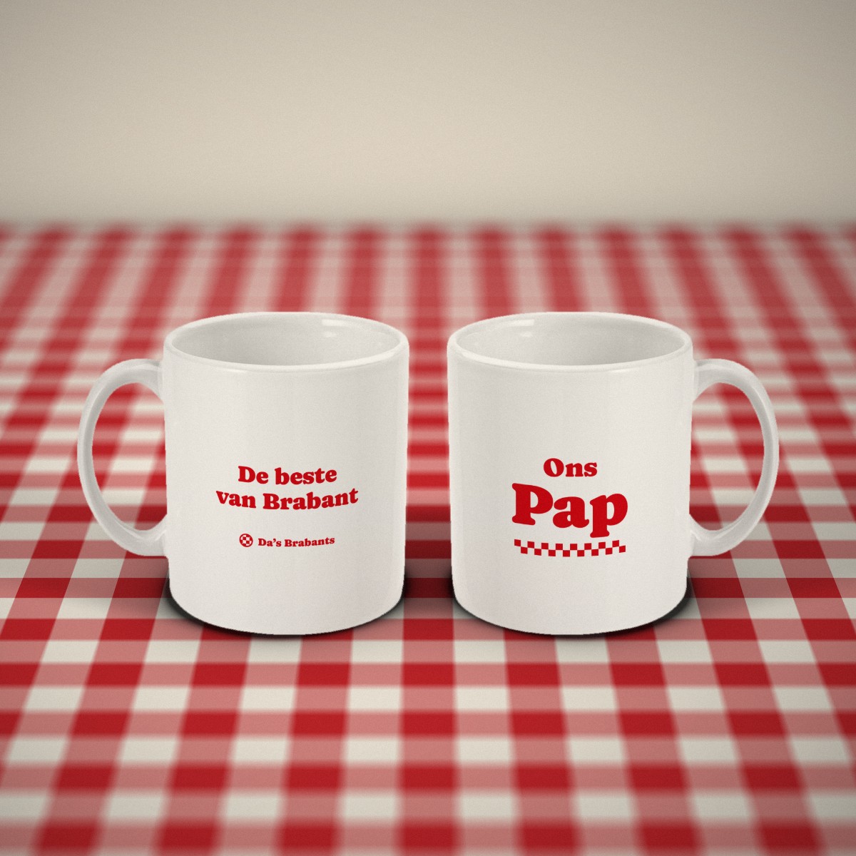 Voor- en achterkant: twee witte mokken „Ons Pap” naast elkaar op een rood-wit geruit tafelkleed