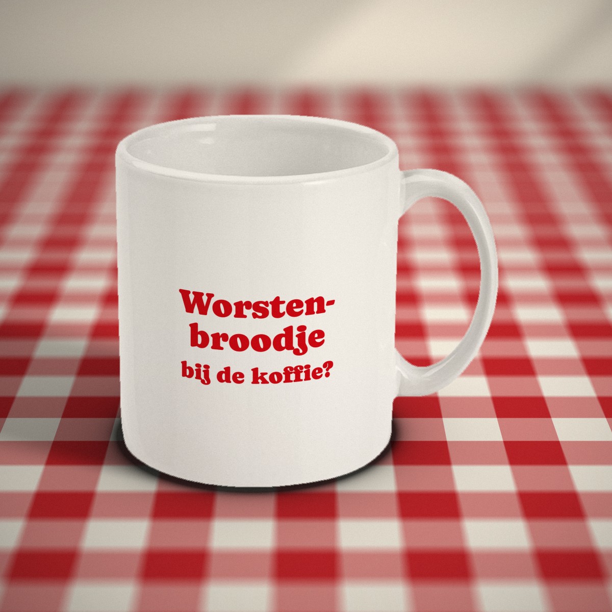 Voorkant: witte mok met in rode letters „Worstenbroodje bij de koffie?”, op een rood-wit geruit tafelkleed
